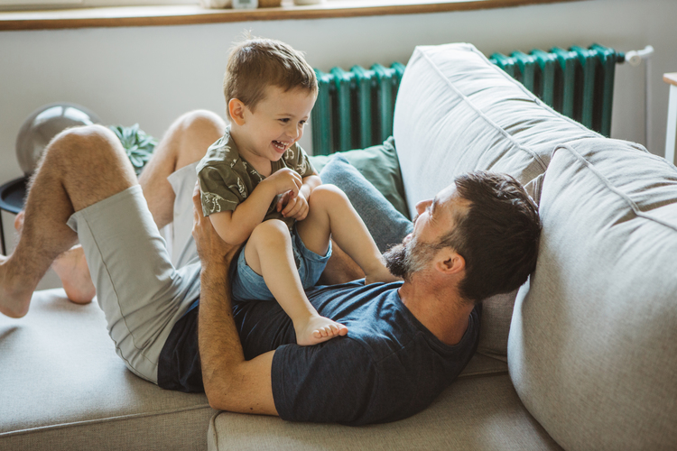 Father lying on a sofa laughing and playing with his smiling young son in a bright living room.