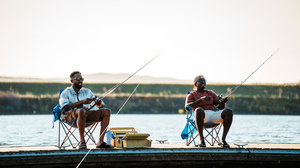 Two smiling men sitting in camping chairs by a river and holding fishing rods.