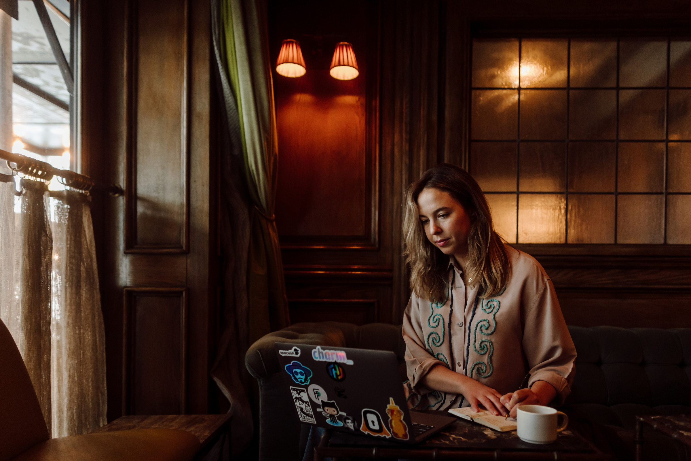 Author Jana sitting with her notebook, computer, and coffee in a dimly-lit room