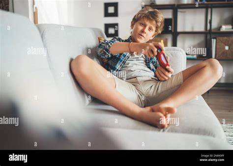 Teenager boy sitting on living room sofa and enthusiastically playing game console using gamepad ...