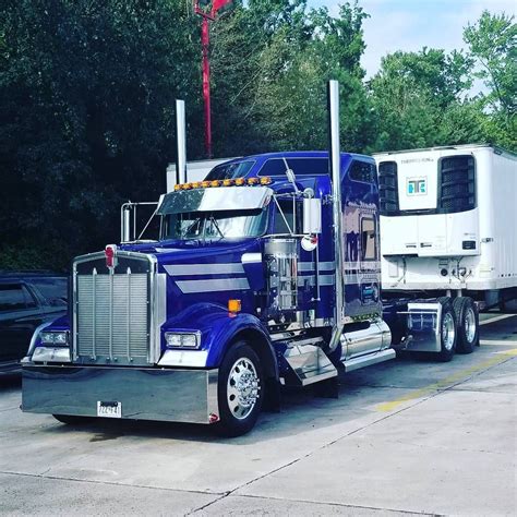 a blue semi truck parked next to a white trailer in a parking lot with trees behind it