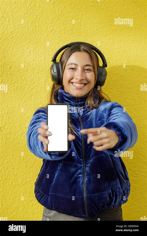 Latin girl with headphones shows the blank screen of her mobile phone isolated on a yellow ...