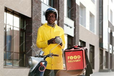 Premium Photo | Young courier in helmet and uniform standing by bike with big red bag