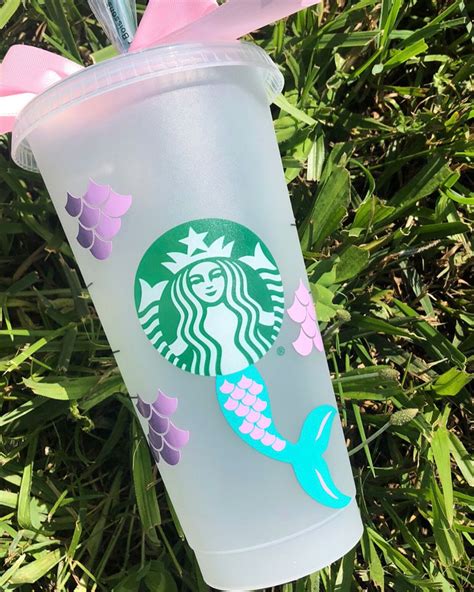 a starbucks cup with a mermaid design on it sitting in the grass next to a pink bow