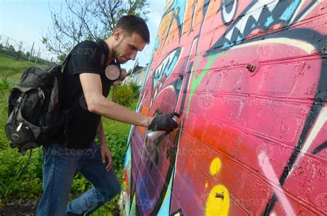 Young graffiti artist with backpack and gas mask on his neck paints colorful graffiti in pink ...