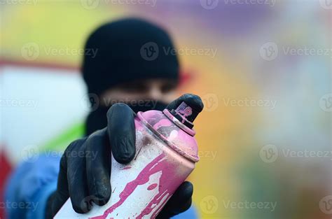 A young graffiti artist in a blue jacket and black mask is holding a can of paint in front of ...