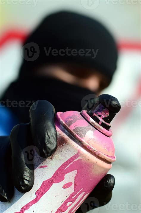 A young graffiti artist in a blue jacket and black mask is holding a can of paint in front of ...