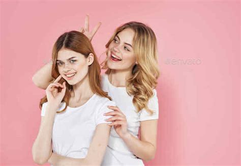 two girlfriends in white t-shirts hugs emotions pink background Stock Photo by shotprime