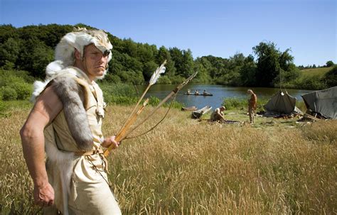 a man dressed in native american garb holding two arrows near a body of water