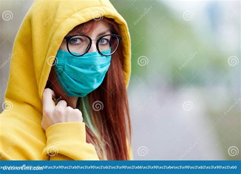 A Young Girl with Glasses, a Reusable Medical Mask, and a Yellow Hood Turns and Looks Around ...