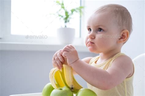 funny baby girl sitting in child chair eating banana on white kitchen ...