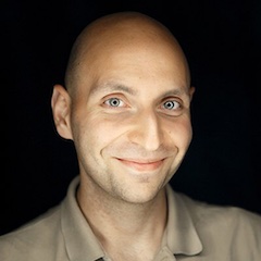 A smiling light-skinned man with a shaved head illuminated in front of a pitch black background.