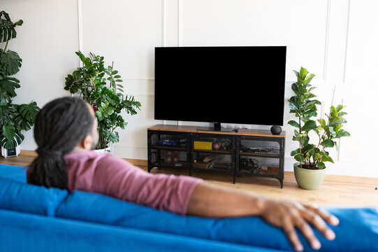 Relaxed African-American man with locks watching TV at home, back view at the TV with a blank black display, mockup, advertising concept