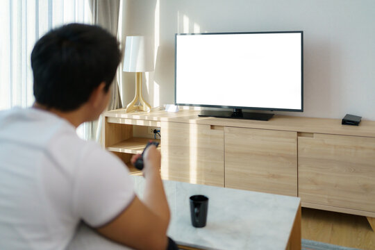 An Asian man holds a TELEVISION remote and is pressing the channel while watching TV on the couch in the living room at home..