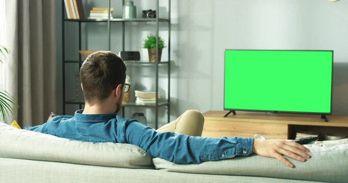 Rear of Caucasian young guy watching TV with chroma key resting on sofa sitting in living room in house. Man watch film on television with green screen sitting on couch in apartment, leisure