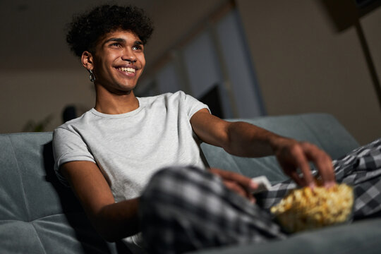 Cheerful young guy wearing pajamas taking some popcorn from a bowl while watching comedy, sitting on a couch at home