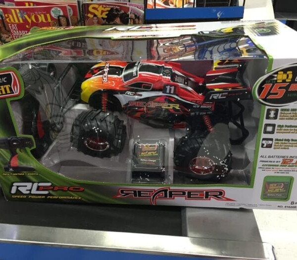 A boxed New Bright R/C Pro Reaper is prominently displayed on the store shelf. This RC car, in bold red, yellow, and black with oversized tires, promises a thrilling 15 mph top speed. The packaging includes a remote controller and indicates it is suitable for ages 8 and up.