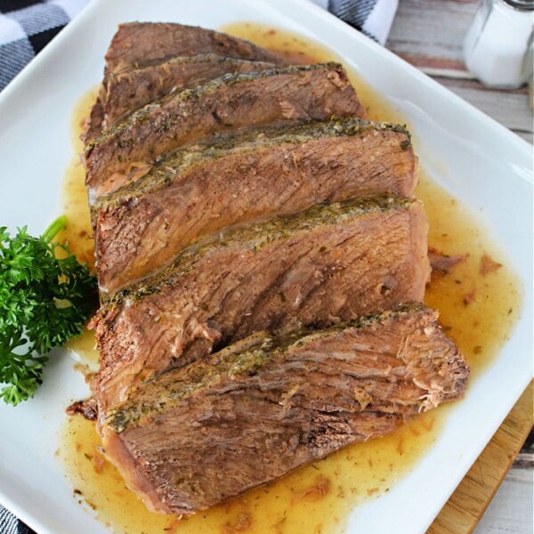 Sliced roast beef on a white rectangular plate, garnished with fresh parsley. A checkered black and white napkin and a salt shaker are placed nearby on a wooden surface. Juices from the frozen roast in the oven are visible on the plate.