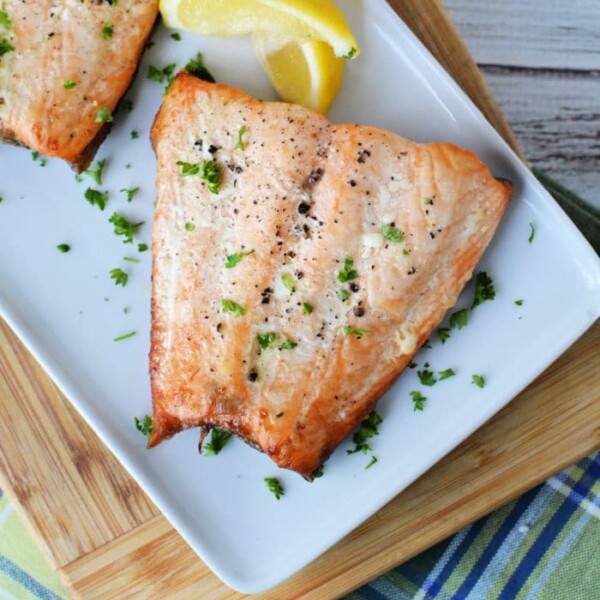 A grilled salmon fillet, previously frozen for freshness, glistens with chopped parsley on a white plate. Lemon wedges accompany it, all elegantly positioned on a wooden cutting board atop a plaid fabric.