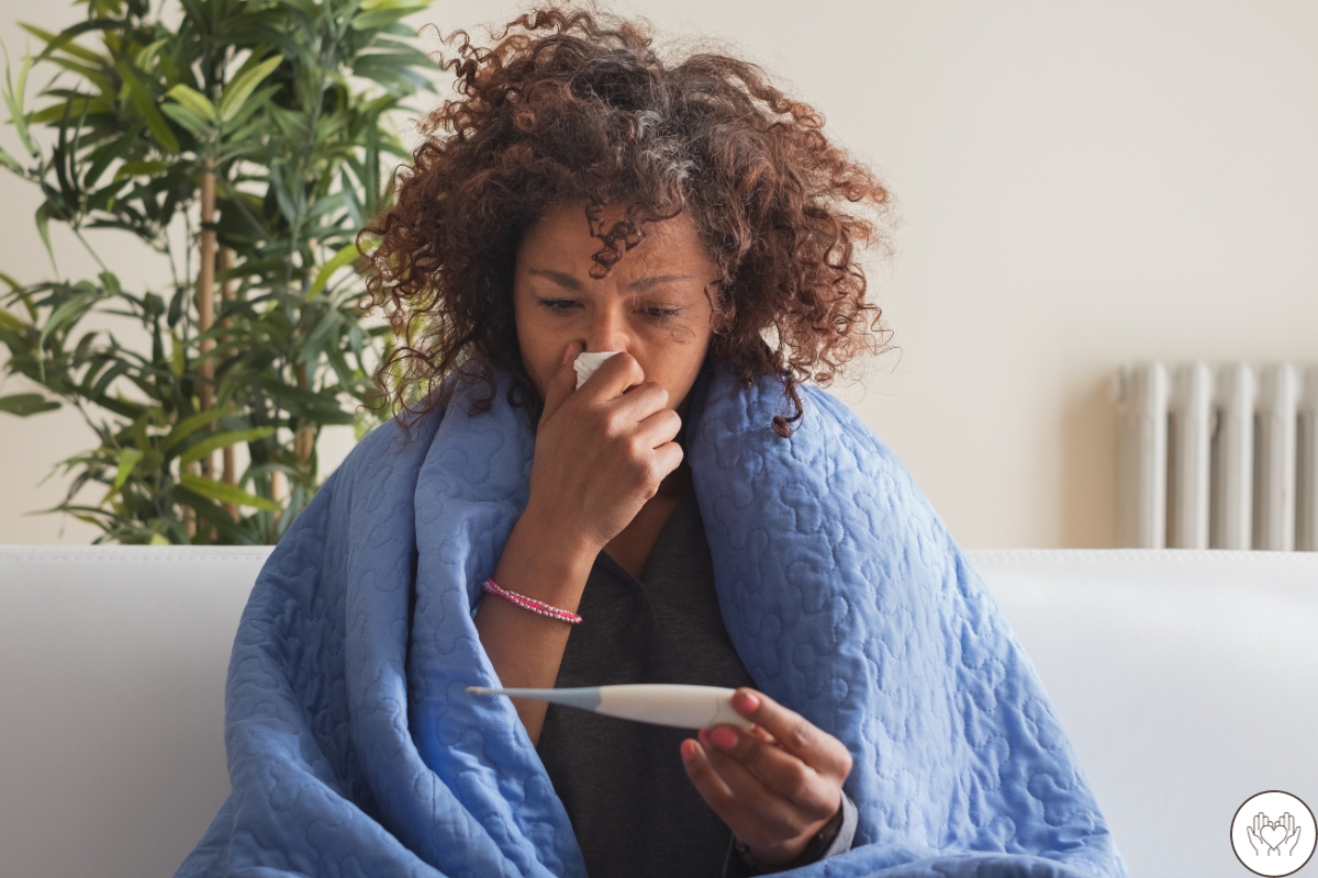 image: woman on couch wrapped in blue blanket looking at thermometer in her hand and holding tissue to her nose with the other.