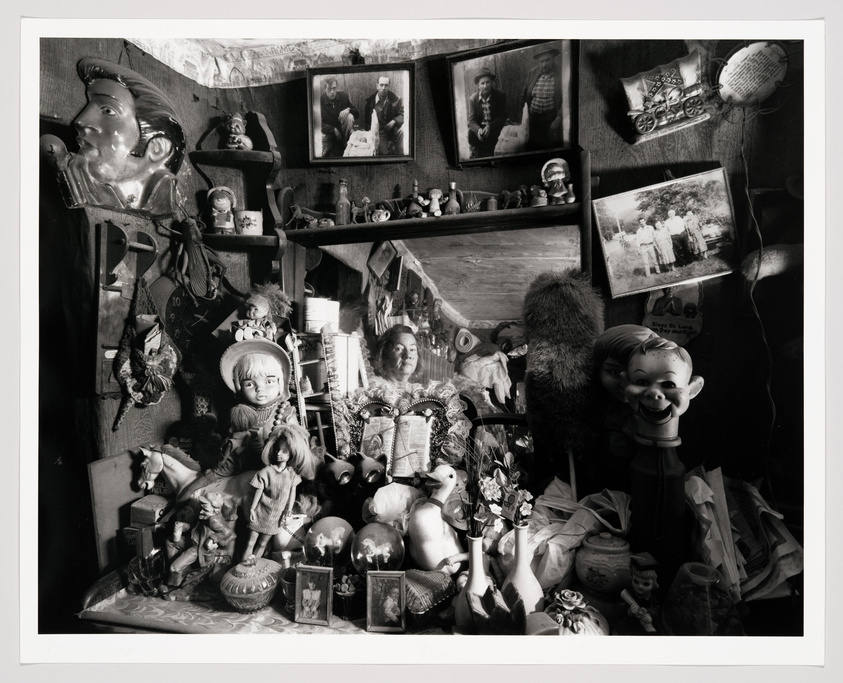 A black and white photograph displaying a densely packed collection of various objects including dolls, figurines, photographs, and paintings. The items are arranged on shelves and the wall, creating a cluttered and eclectic display. Some of the notable objects include a large doll head, a figurine of a boy with a big smile, and several framed pictures of people. The overall impression is one of a personal and nostalgic assemblage.