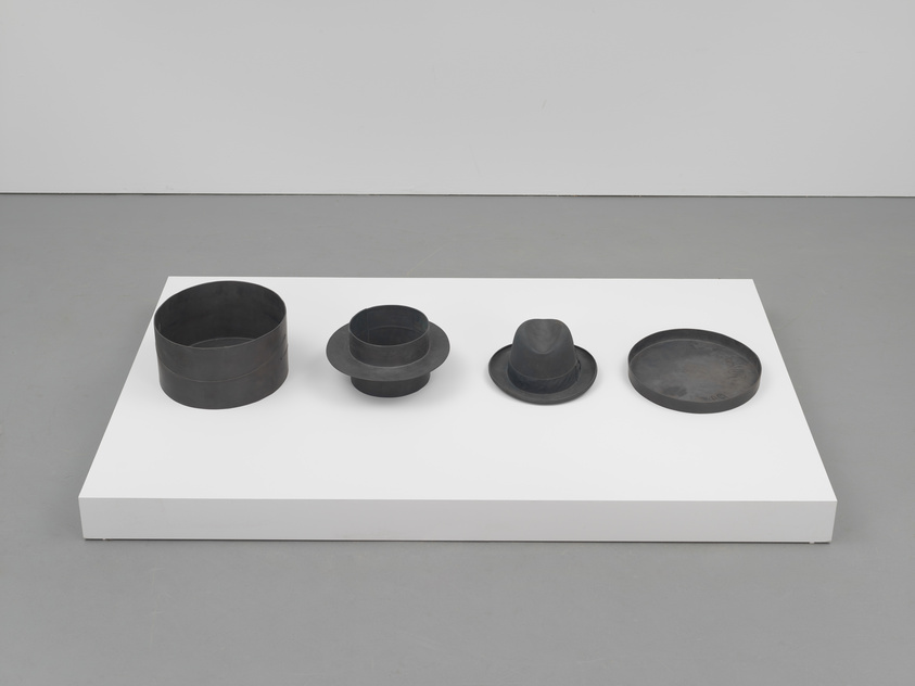 A modern art installation featuring four black, cylindrical objects of varying sizes and shapes arranged in a line on a white rectangular platform against a grey floor and white wall background. The second object from the left resembles a hat with a brim.