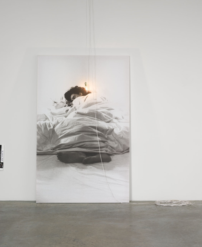 A large monochromatic photograph of a person lying in bed is displayed in a gallery setting, with the image extending onto the floor where a cable seems to connect the photograph to a power source.