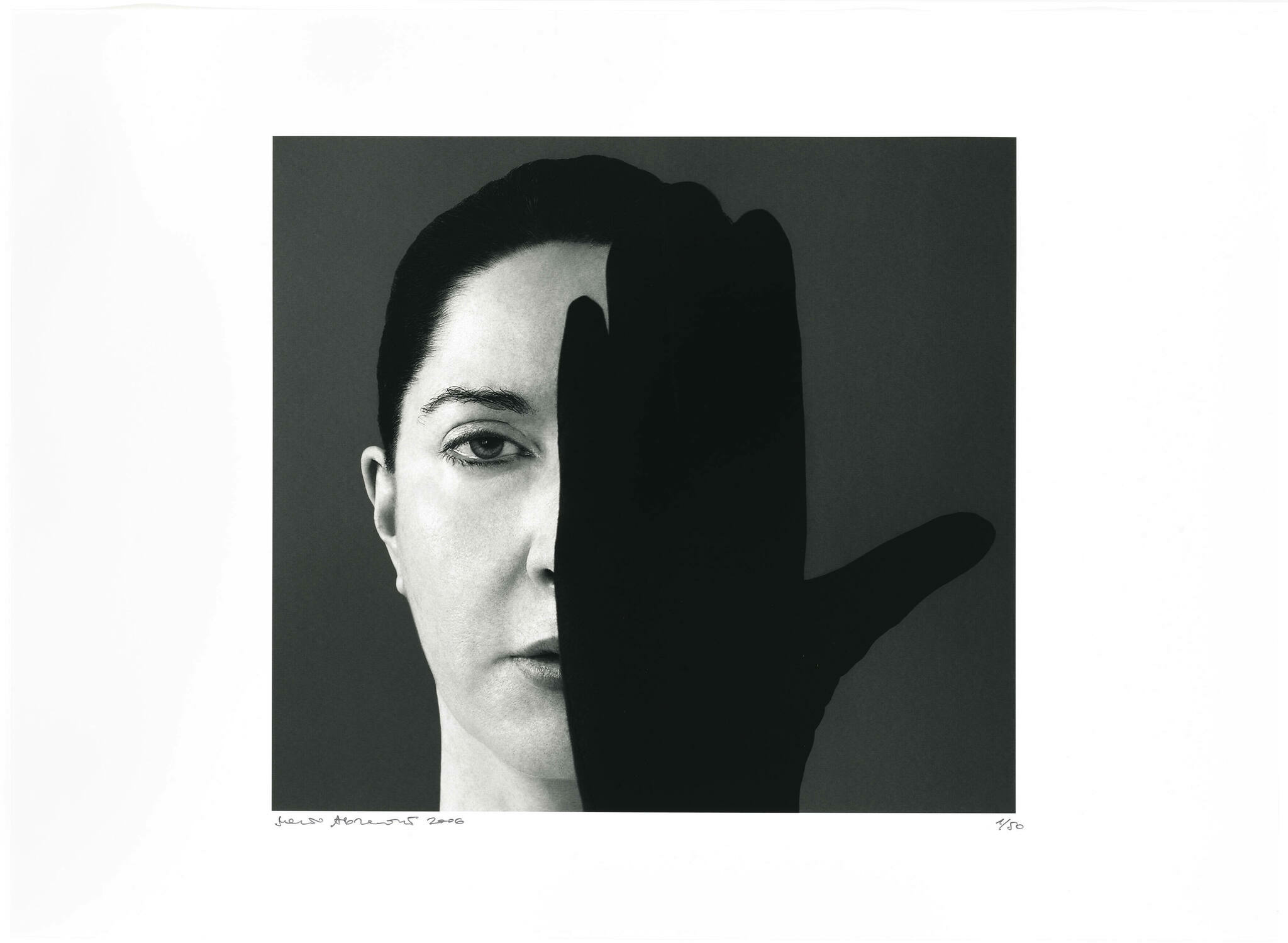 Black and white photograph of a woman's face partially obscured by a silhouette of a hand in the foreground. The image has a stark contrast between the light and dark areas, with the woman's expression appearing neutral. There is text at the bottom left of the image, likely indicating the artist's signature and the year, and a fraction at the bottom right, possibly denoting the print number of the artwork.