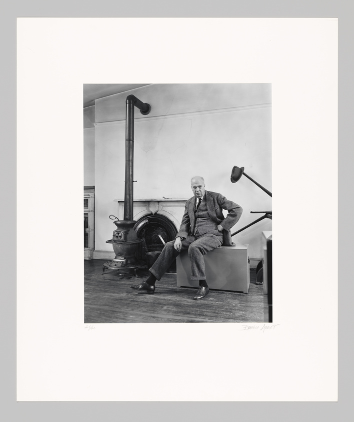 Black and white photo of a seated elderly man in a suit, by a potbelly stove and angled desk lamp, in a room with plain walls.