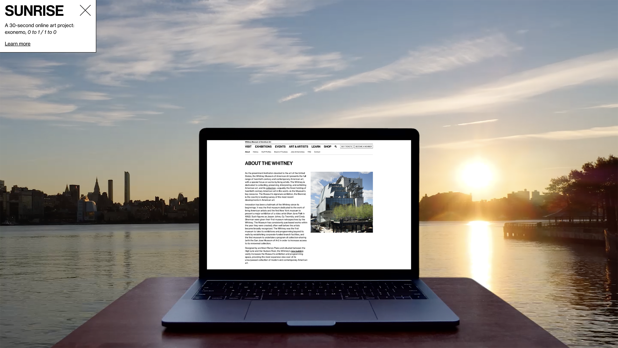 A laptop with whitney.org onscreen sits on a table by the water as the sun rises over Manhattan.