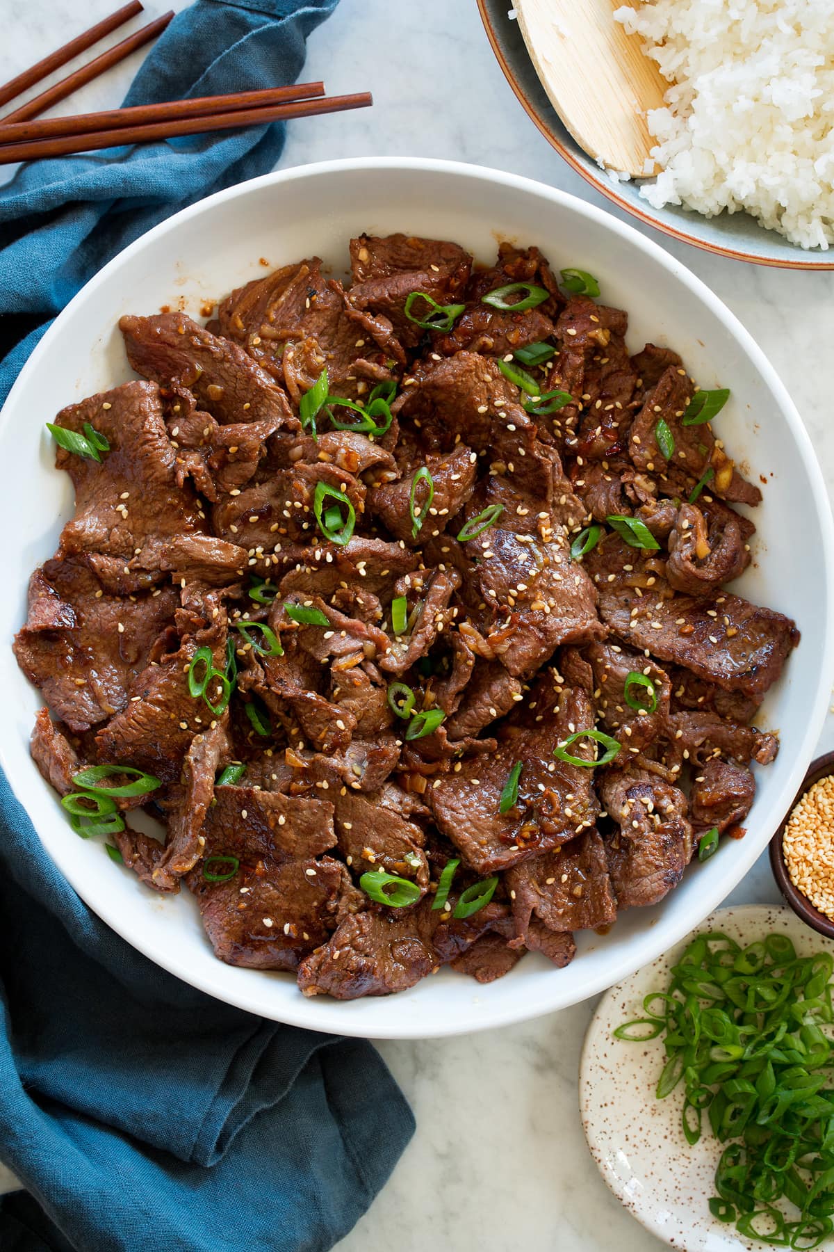 Bulgogi shown overhead in a white serving bowl with wooden chopsticks, rice, green onions and a blue cloth to the side.