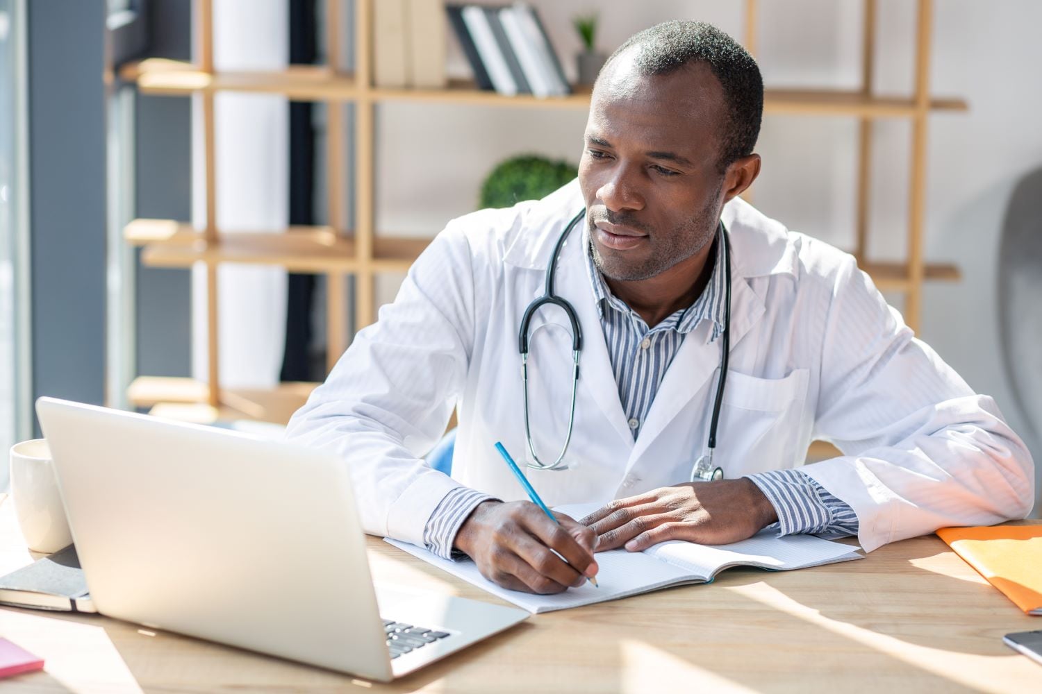 A doctor looking at his computer while taking notes at his desk.