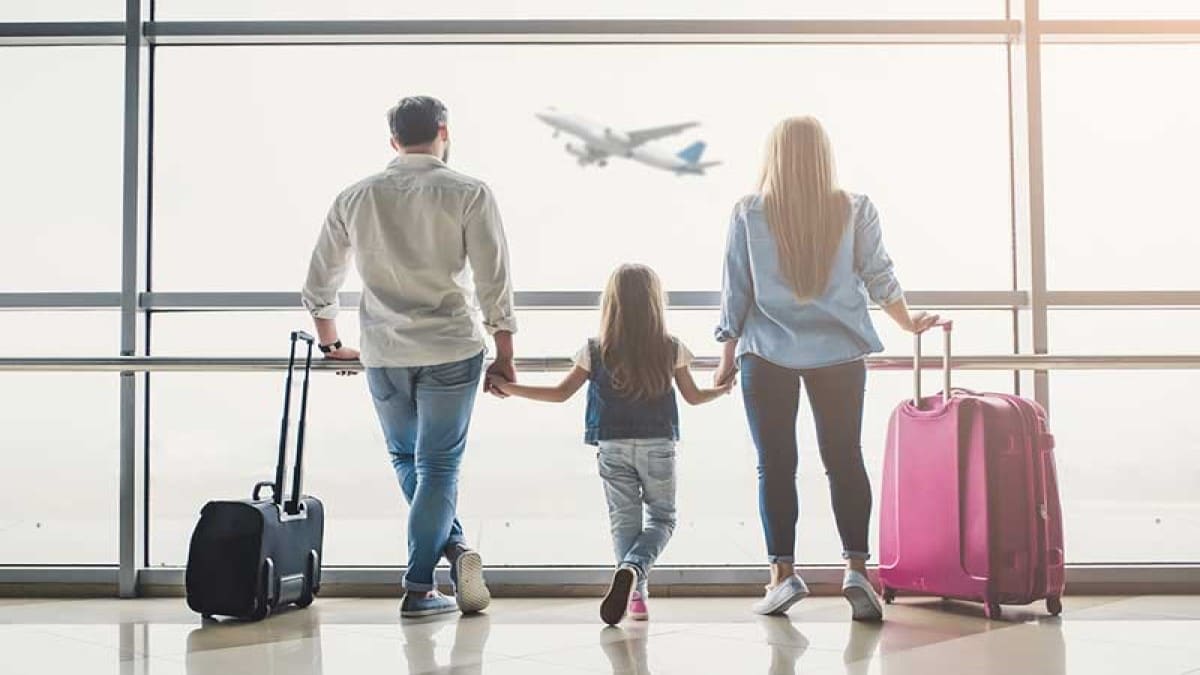 Three people with suitcases looking at a plane in the sky.