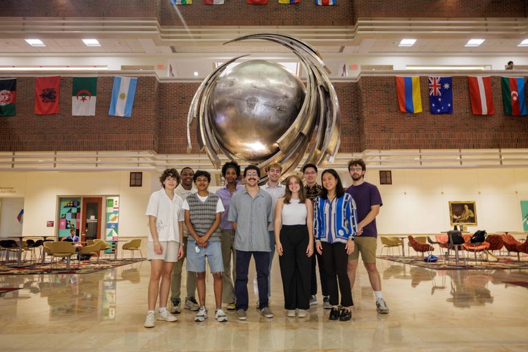 Cohort of students posed in front of a large metal statue in a building atrium