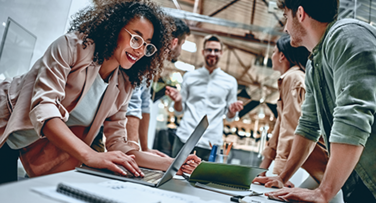 Some people stand together leaning on a desk, a woman operates a laptop