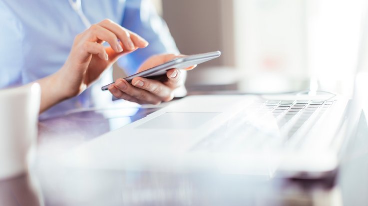 A person sits in front of an opened laptop and operates a smartphone