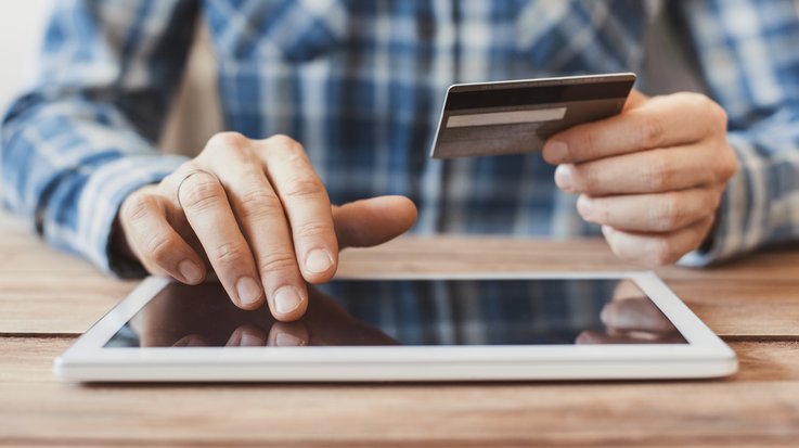 A man sits at a table and operates a tablet while holding a credit card in one hand