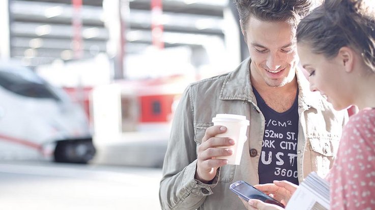 Two young people standing in front of a train station operating a smartphone