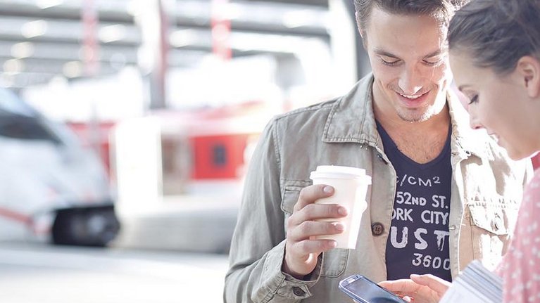 Two young people standing in front of a train station operating a smartphone