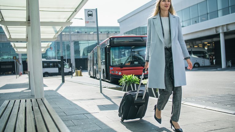 Eine Geschäftsfrau an einem Busterminal, mit einem Rollkoffer, den sie hinter sich her zieht