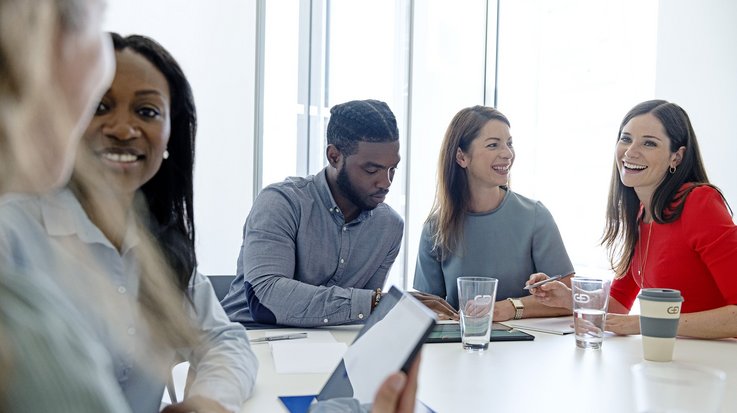 Some employees sit at a large table in a meeting room and talk to each other