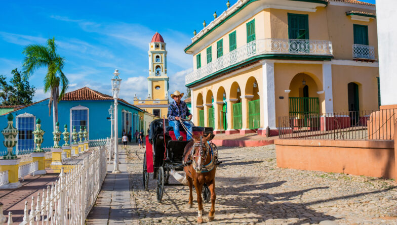 A horse-drawn carriage moves slowly along a cobbled street in Trinidad, Cuba, surrounded by colonial buildings and swinging palm trees