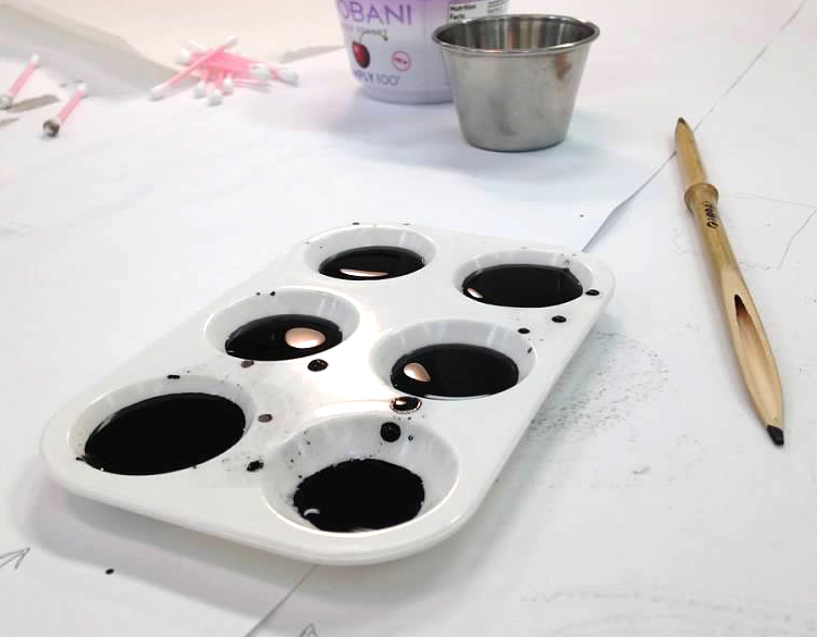 A tray with black ink, a wooden brush, and a metal cup are on a table, possibly used for drawing classes.