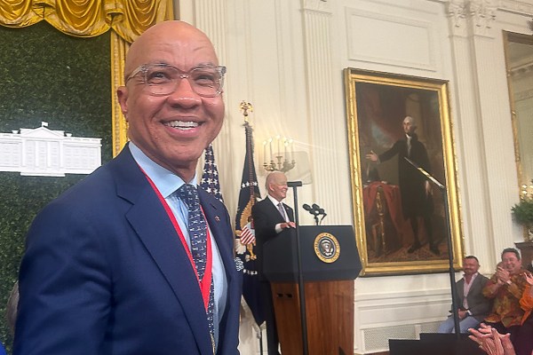 A man in a suit, Darren Walker, smiles at the camera in a room with a podium displaying the U.S. presidential seal. Another person, President Biden, is speaking at the podium, with portraits and ornate curtains in the background.