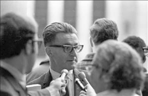 Koome wearing thick rim glasses, combed back hair and a suit, being interviewed by at least three reporters holding microphones