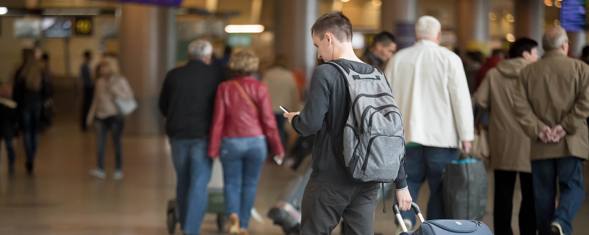 A man using his phone while rolling a suitcase at a  transportation center 