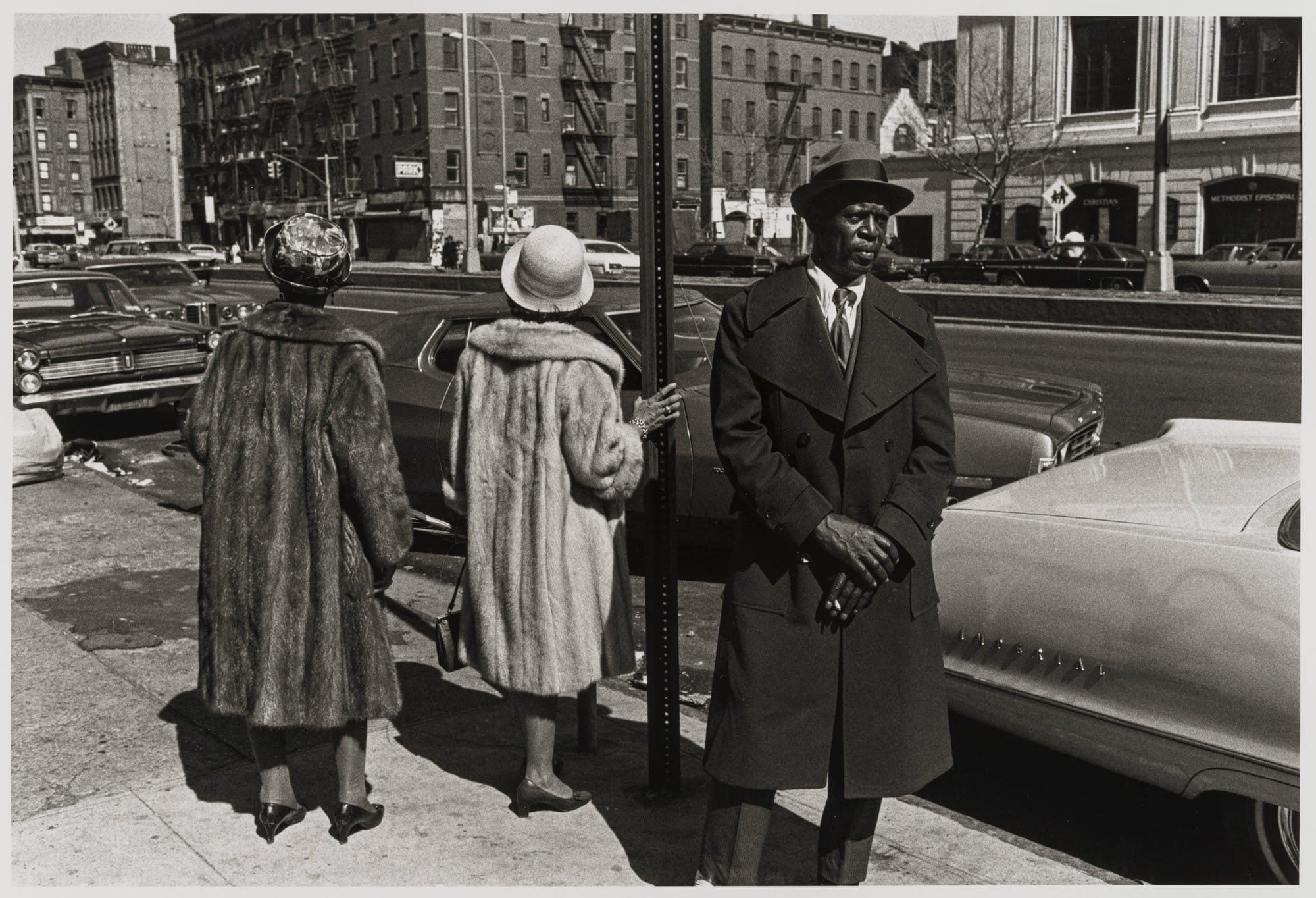 a black-and-white photo by Dawoud Bey of a Black man standing on a street in New York next to two women in large coats and hats, who are turned away from the camera