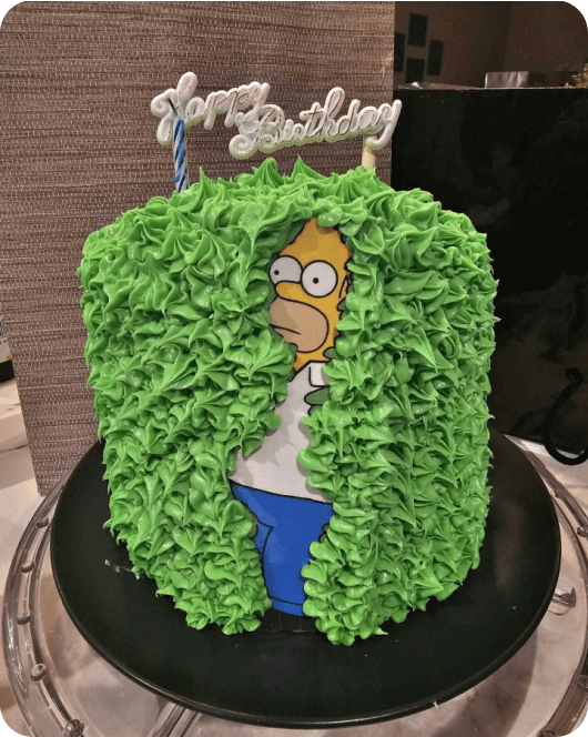 Birthday cake with Homer Simpson sinking into green icing replicating a famous meme of Homer disappearing into a hedge