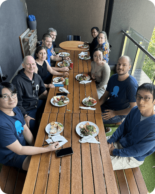 Ten people sitting outside at wooden tables eating lunch