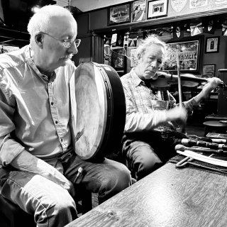 A man holding a bodhrán next to a man playing fiddle at a pub table.